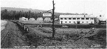Historic black-and-white photo of a sawmill building with a sign and cabins in the background.