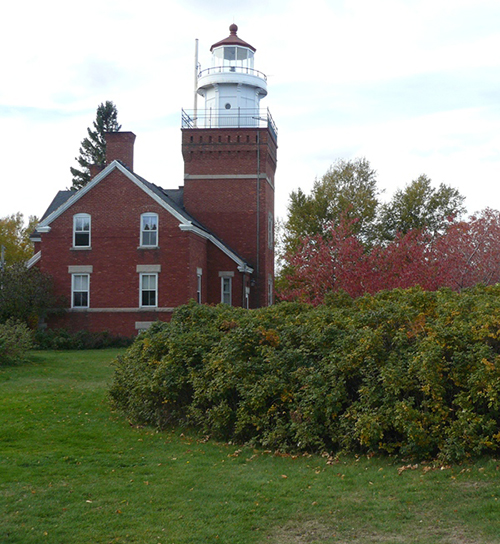 Red-brick lighthouse tower and house, surrounded by green lawn and autumn foliage.