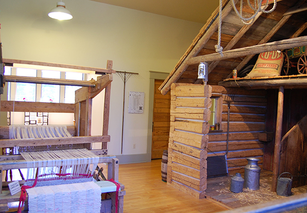 Historical exhibit with a wooden loom and a log cabin interior.