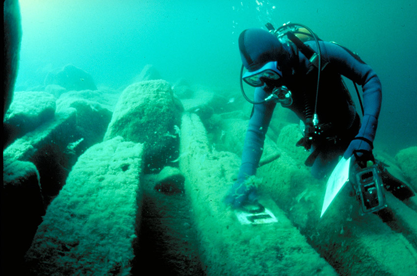 Diver in scuba gear brushes algae from submerged timbers with a handheld device.
