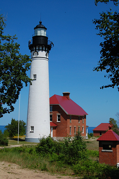 Tall white lighthouse with red-roofed brick buildings under a blue sky.