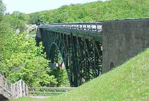 Green cantilever deck bridge spans a lush forested gorge, with stone abutments.
