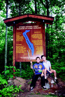 Two kids posing in front of a wooden sign for Alligator Eye Scenic Overlook in a forest.