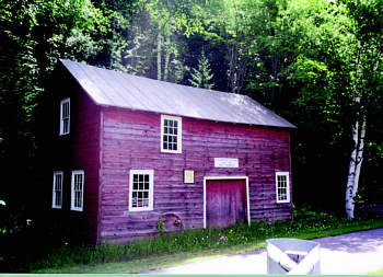 Historic red wooden blacksmith shop with a pitched roof, surrounded by lush green trees.