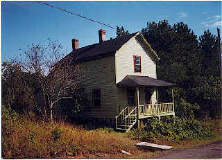 Two-story historic wooden house with small porch, set amid overgrown vegetation.