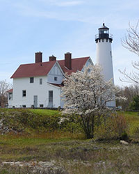 White lighthouse with red-roofed keeper’s house and blooming tree at Pt. Iroquois Historic Site.
