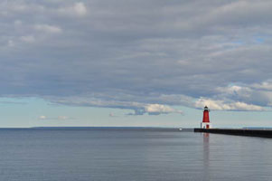 Red lighthouse at the end of a pier under a cloudy sky on calm water.