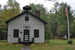 Small white one-room schoolhouse with bell tower and flagpole surrounded by trees.