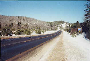 Snowy roadside along US 45 with forested hills in the background and a historical marker ahead.