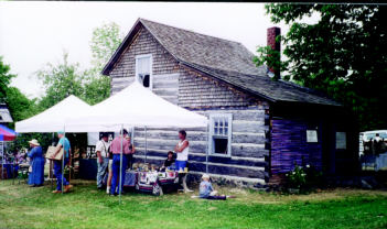 Visitors browse crafts outside a restored log cabin at an early mining village heritage site.