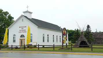 White mission-style building labeled "Museum of Ojibwa Culture" with a teepee and sign nearby.