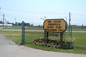  Sign at entrance to Upper Peninsula State Fairgrounds in Escanaba, with flowers and fenced field.